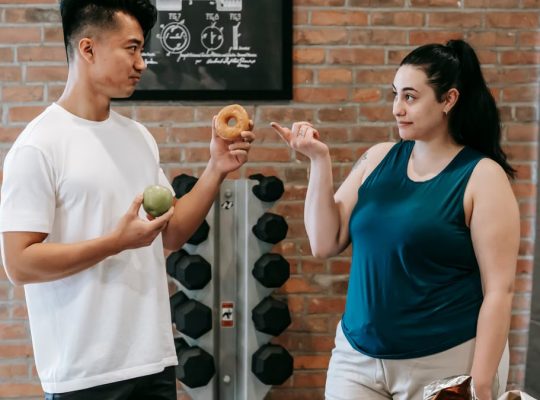 Side view of Asian personal instructor with apple and doughnut offering healthy food to plus size woman while standing in gym