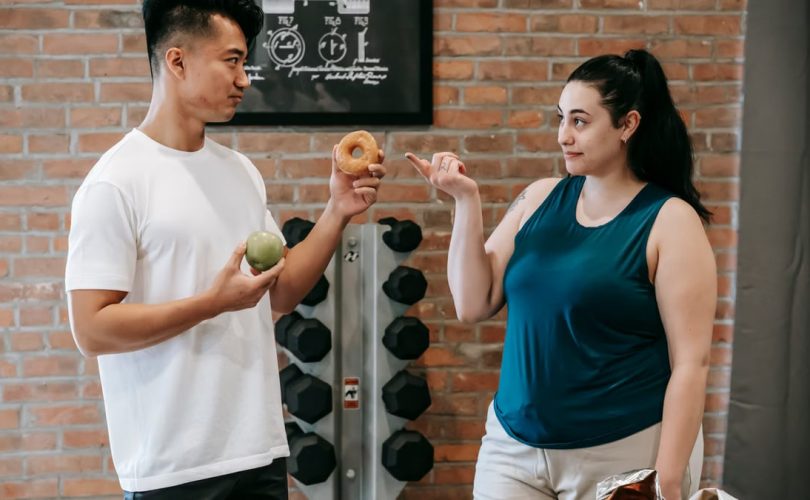 Side view of Asian personal instructor with apple and doughnut offering healthy food to plus size woman while standing in gym